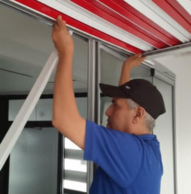 عَامِل يَقُوم بِتَرْكِيب سَقْف أَوْ مِظَلَّة مِنْ الأَلُومِنْيُوم بِخَطُوط حَمْرَاء وَبَيْضَاء دَاخِلَ مَكَان مَا.