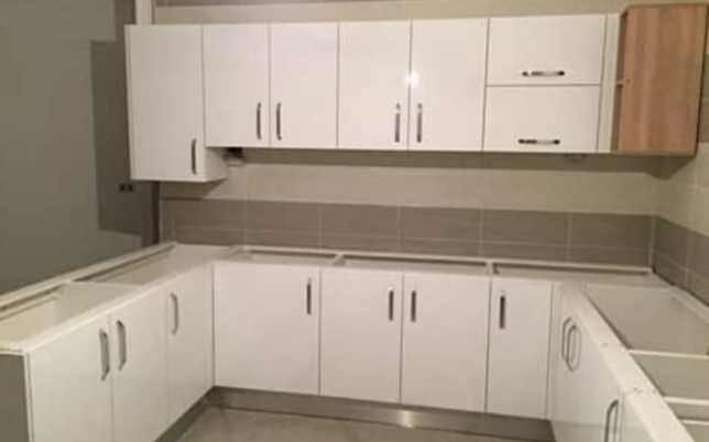 خزائن مطبخ MDF بيضاء لامعة على شكل حرف U مع مقابض فضية، وتظهر وحدة علوية بلون الخشب.